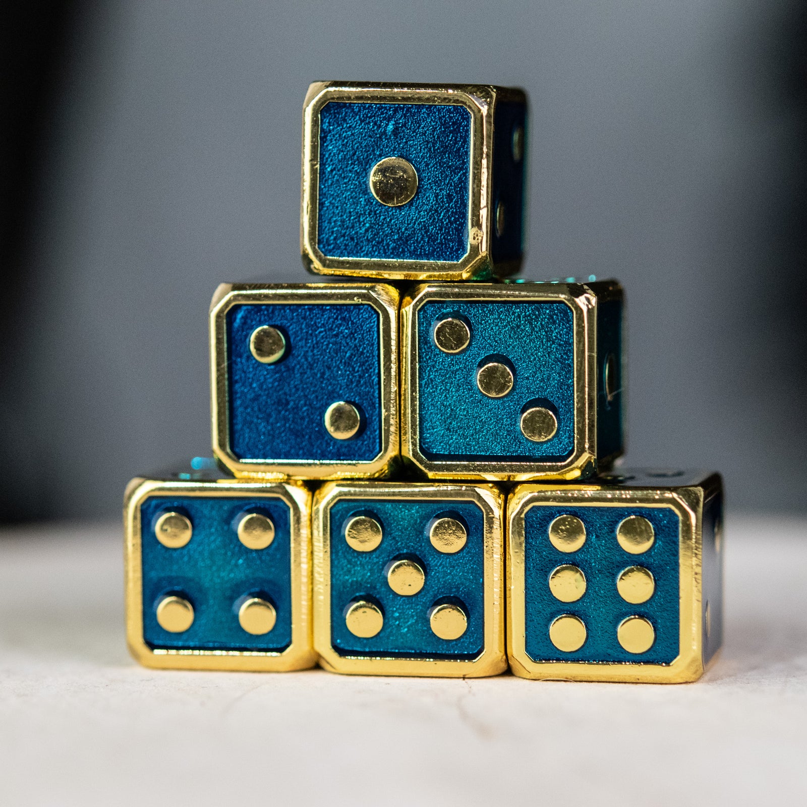 A stack of six metal d6 dice with aqua blue and gold colors, embossed with pips 