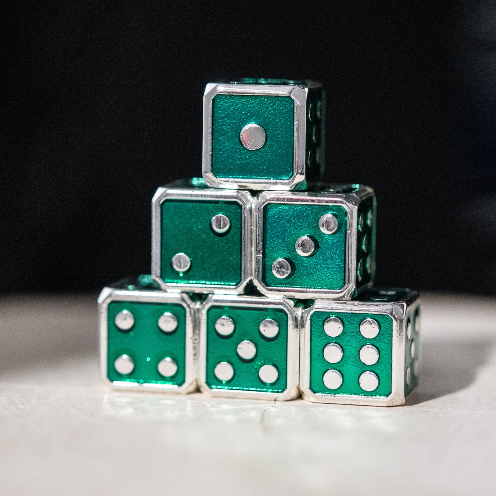 A stack of six green metal d6 dice with silver pips, arranged to form a pyramid shape.
