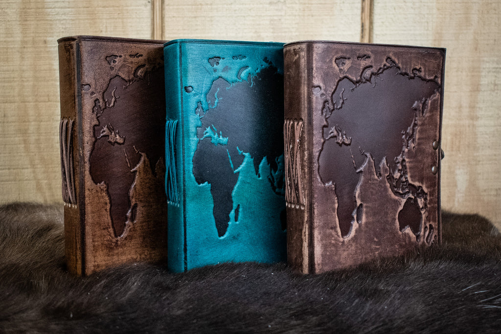 Three leather journals featuring the world map in light brown, blue, and dark brown
