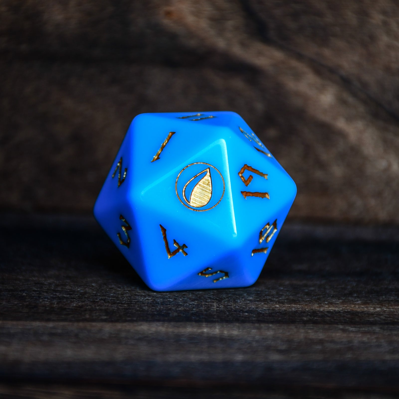 A blue acrylic D20 dice with engraved numbers and a spindown design, placed on a wooden surface.