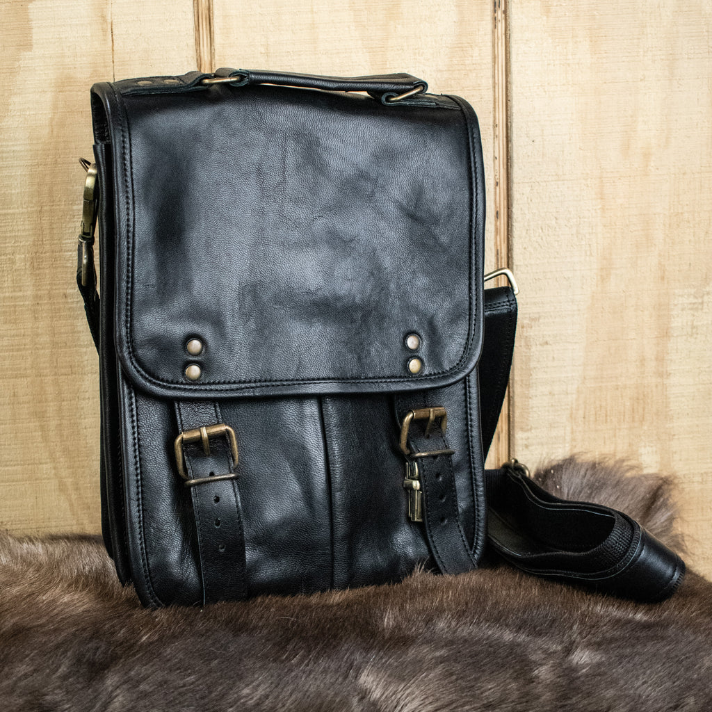 A black full-grain goat leather bag with brass buckles and an adjustable strap, displayed against a wooden background.