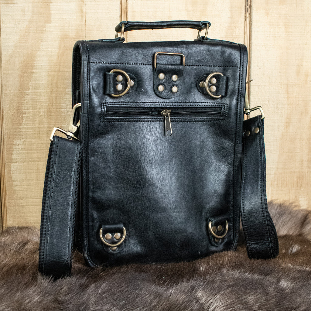 A black full-grain goat leather bag with brass buckles and an adjustable strap, displayed against a wooden background.