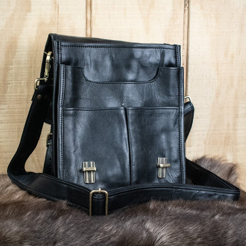 A black full-grain goat leather bag with brass buckles and an adjustable strap, displayed against a wooden background.