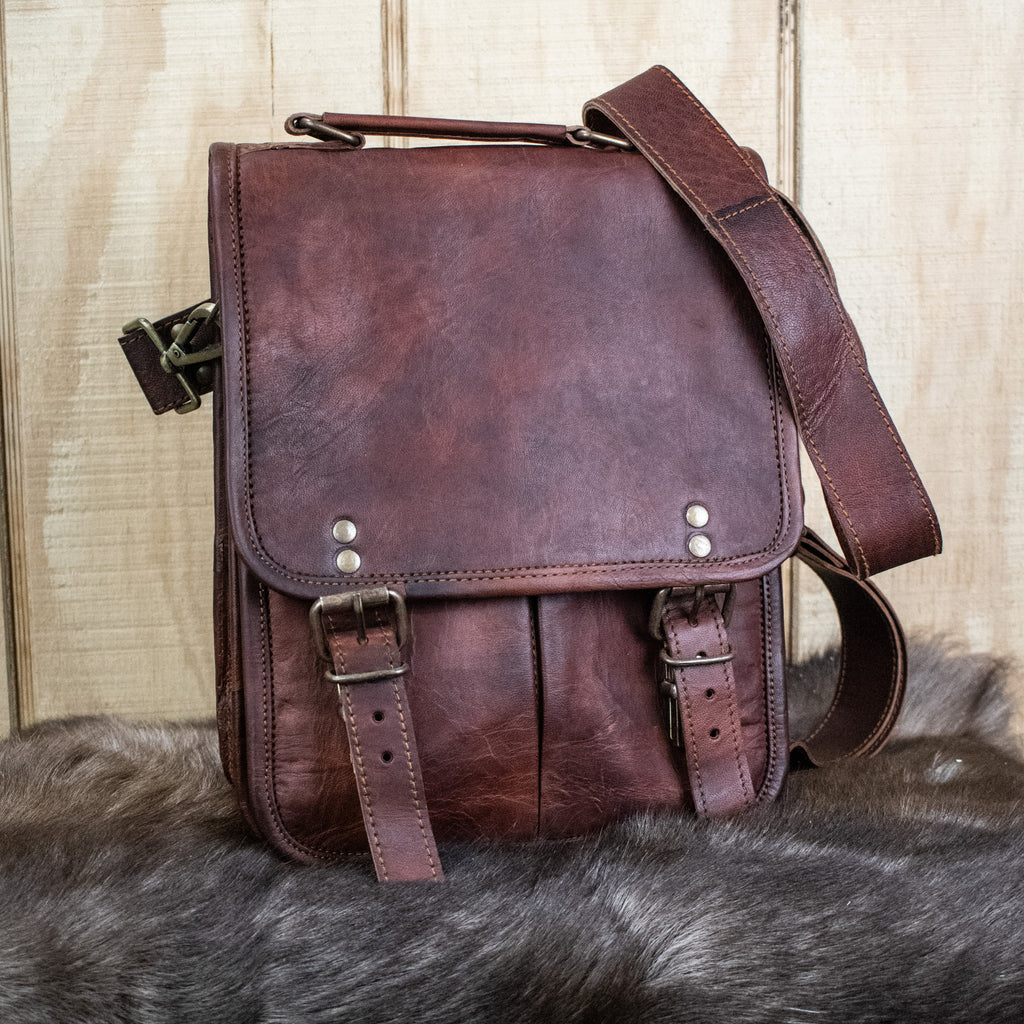 A brown full-grain leather bag with brass buckles, an adjustable strap, and pockets, displayed on a furry surface.