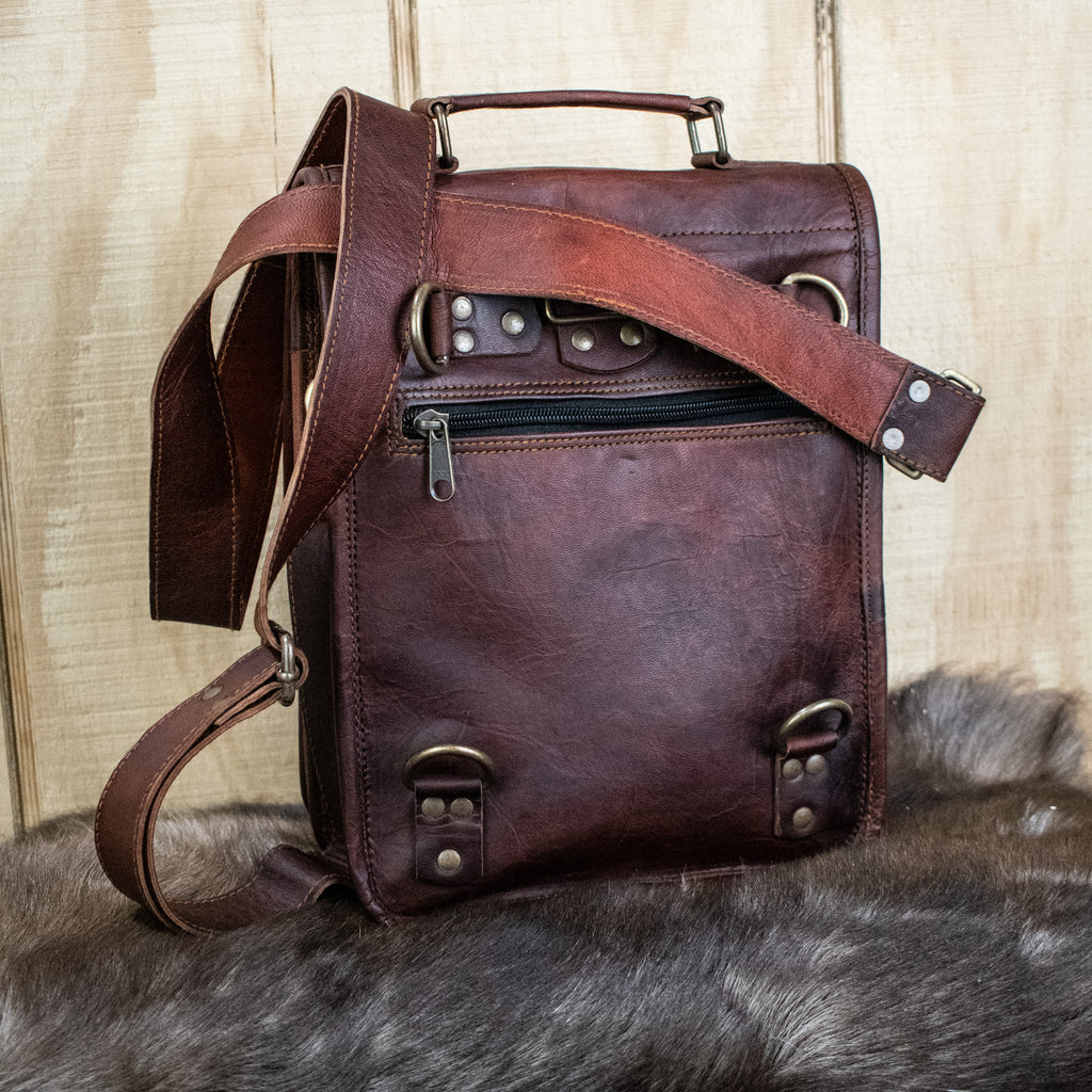 A brown full-grain leather bag with brass buckles, an adjustable strap, and pockets, displayed on a furry surface.
