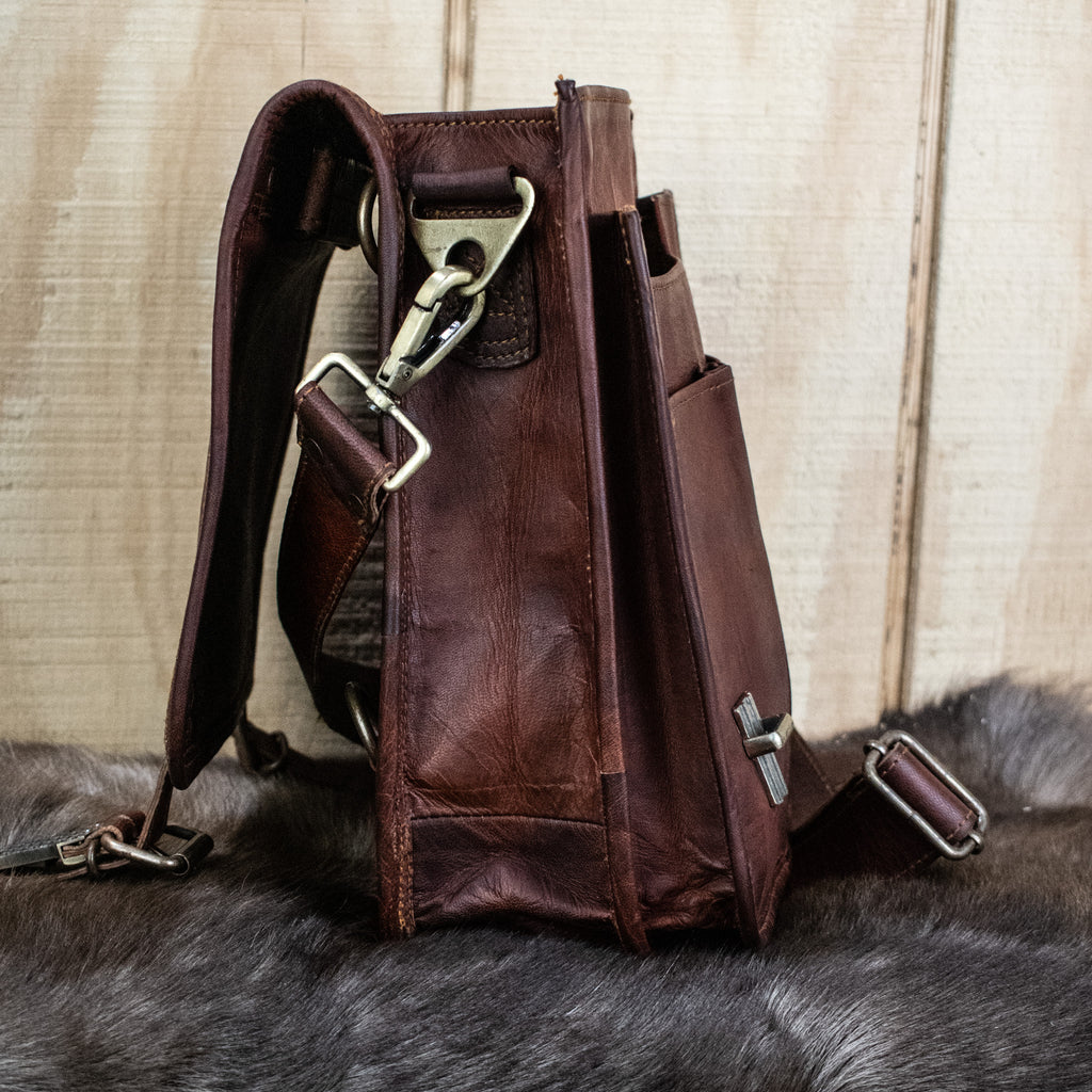A brown full-grain leather bag with brass buckles, an adjustable strap, and pockets, displayed on a furry surface.