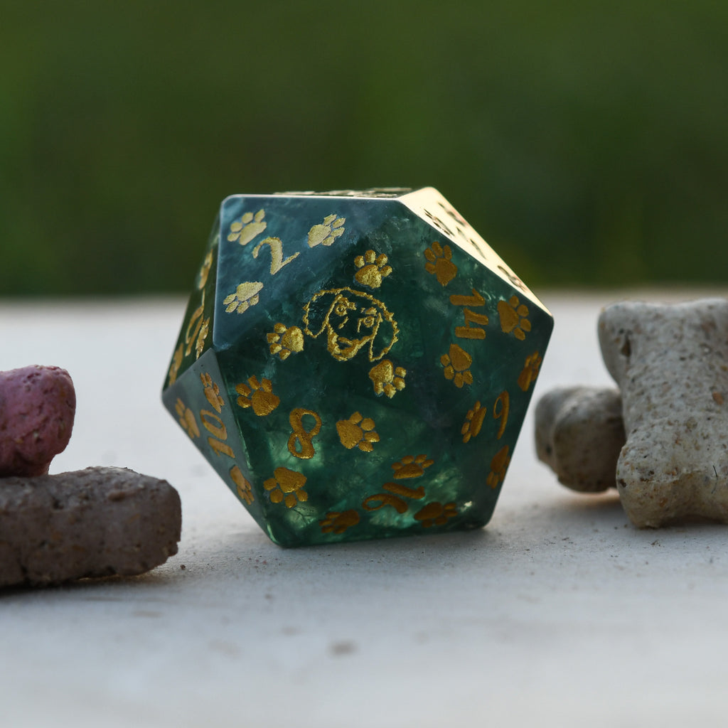 A Dachshund Puppy Paws Green Fluorite 30mm D20 with gold paint details, shaped as a D20, displayed on a natural stone surface.