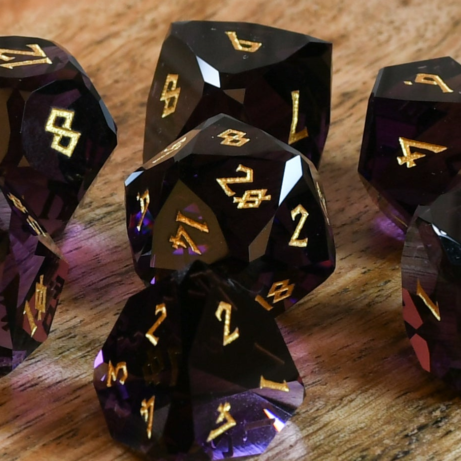 A set of multifaceted glass dice in deep plum color with gold engraved numbers, displayed on a wooden surface.
