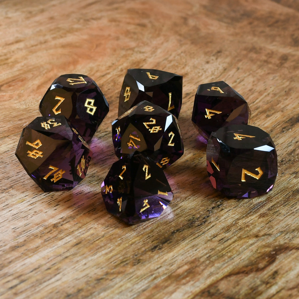 A set of multifaceted glass dice in deep plum color with gold engraved numbers, displayed on a wooden surface.