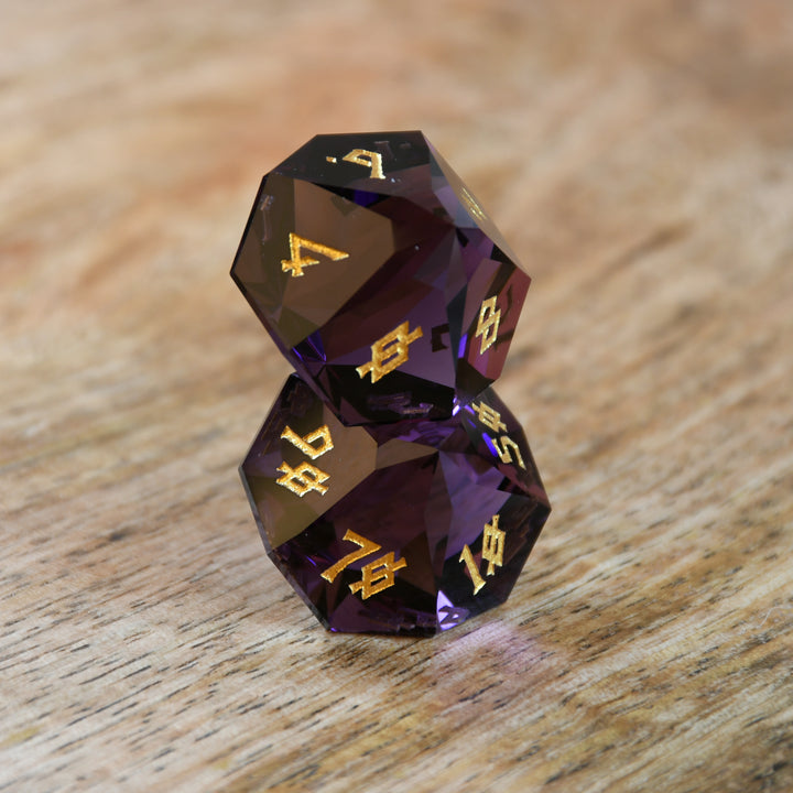 A set of multifaceted glass dice in deep plum color with gold engraved numbers, displayed on a wooden surface.