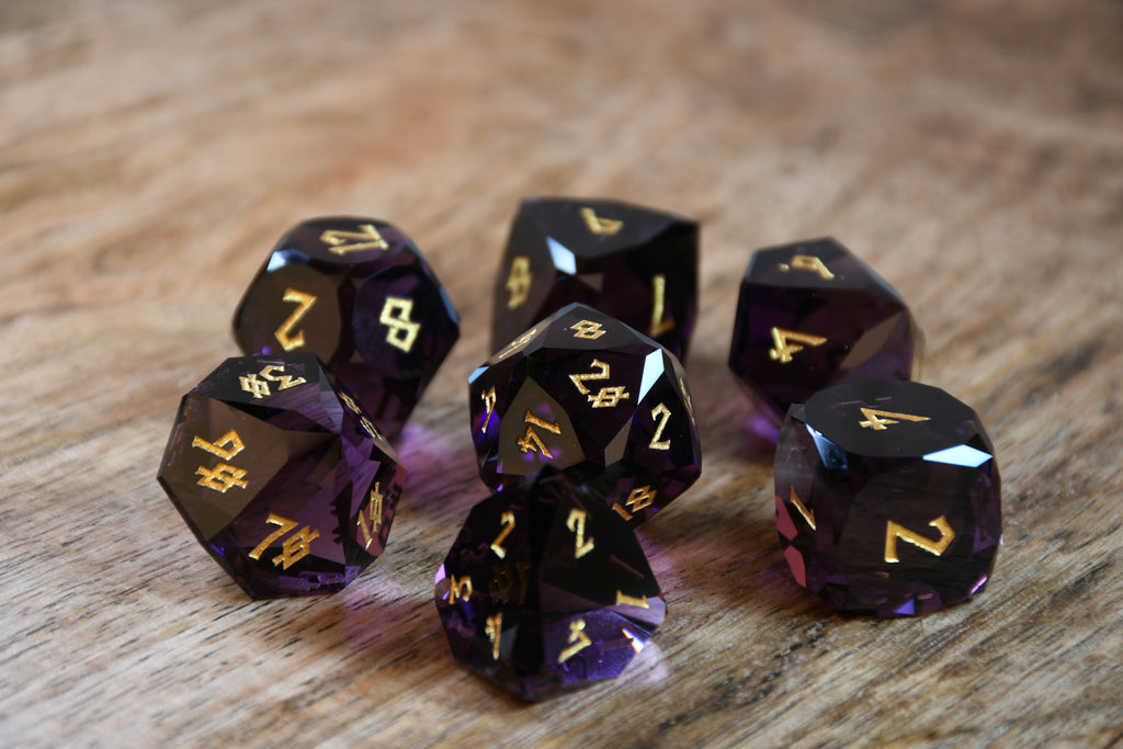 A set of multifaceted glass dice in deep plum color with gold engraved numbers, displayed on a wooden surface.
