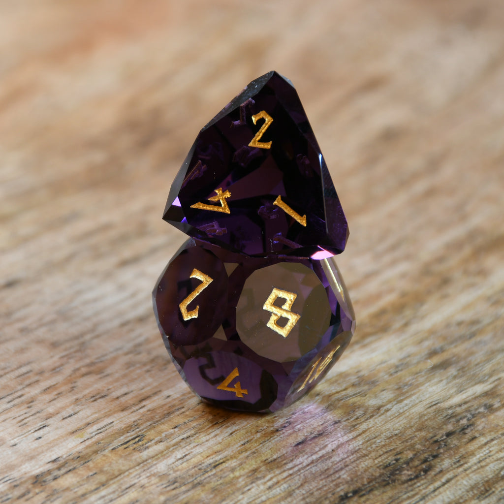 A set of multifaceted glass dice in deep plum color with gold engraved numbers, displayed on a wooden surface.