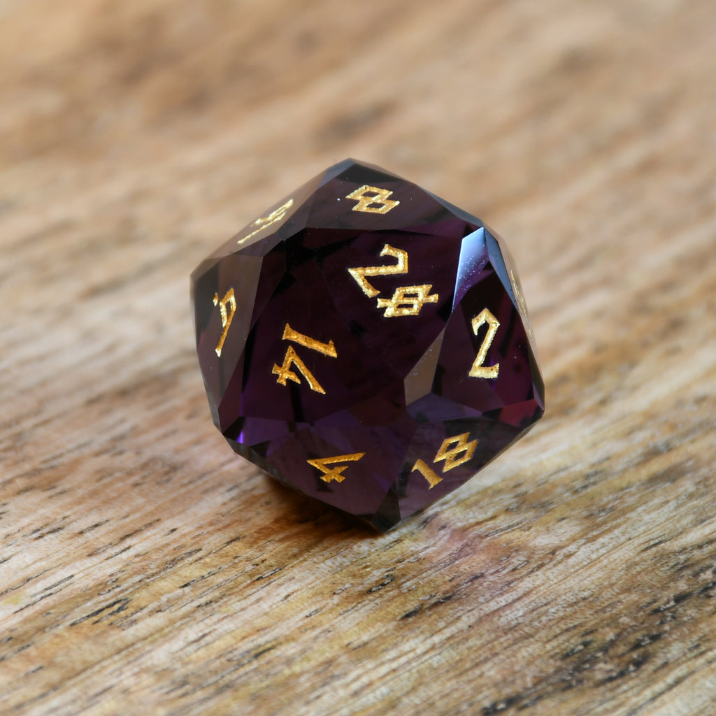 A set of multifaceted glass dice in deep plum color with gold engraved numbers, displayed on a wooden surface.