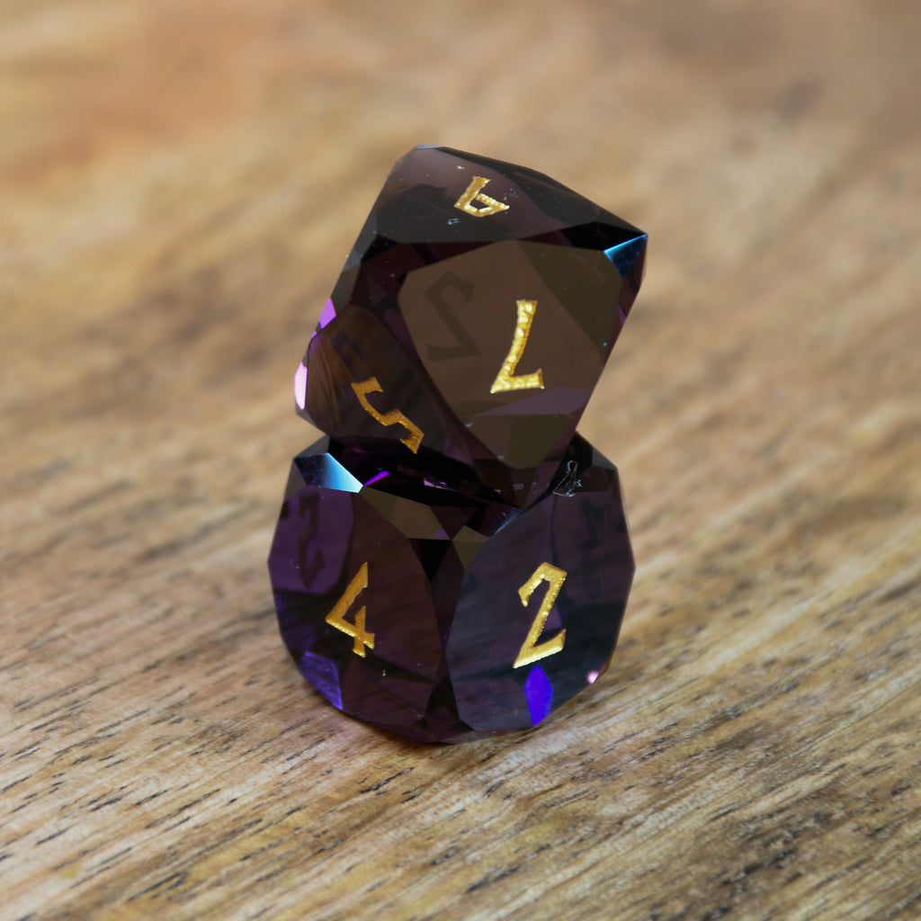 A set of multifaceted glass dice in deep plum color with gold engraved numbers, displayed on a wooden surface.
