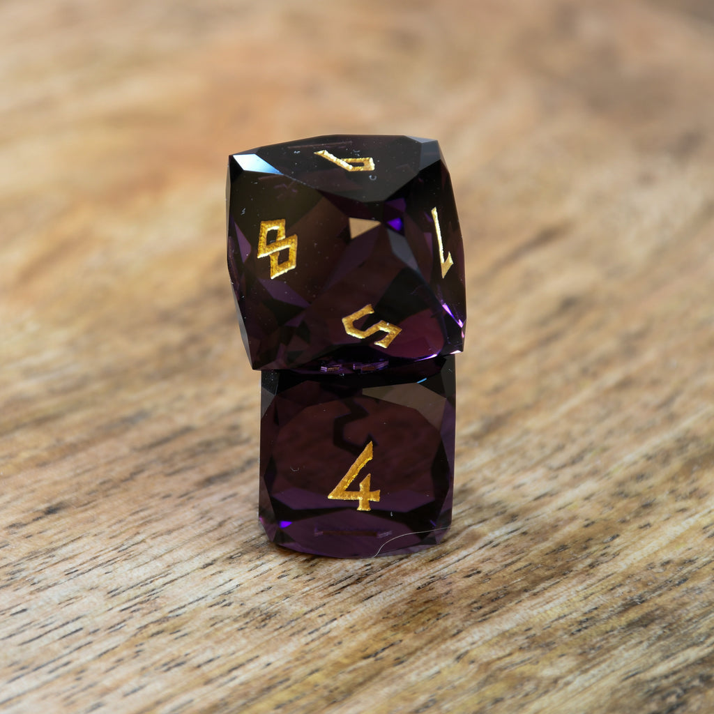 A set of multifaceted glass dice in deep plum color with gold engraved numbers, displayed on a wooden surface.