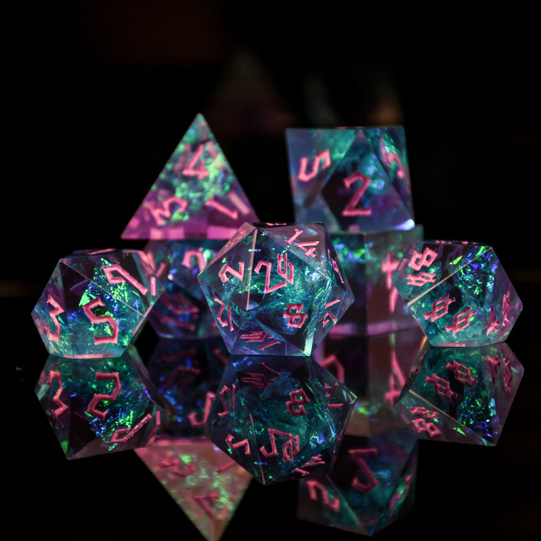 A set of sharp-edged, multicolored resin dnd dice with pink hand-painted numbers, reflecting a vibrant blue, green and purple glow.
