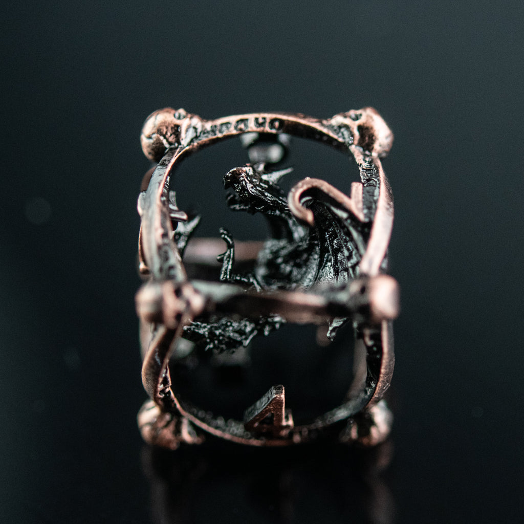 A set of bronze hollow metal dice with embossed numbers and dragons inside, displayed on a reflective surface.