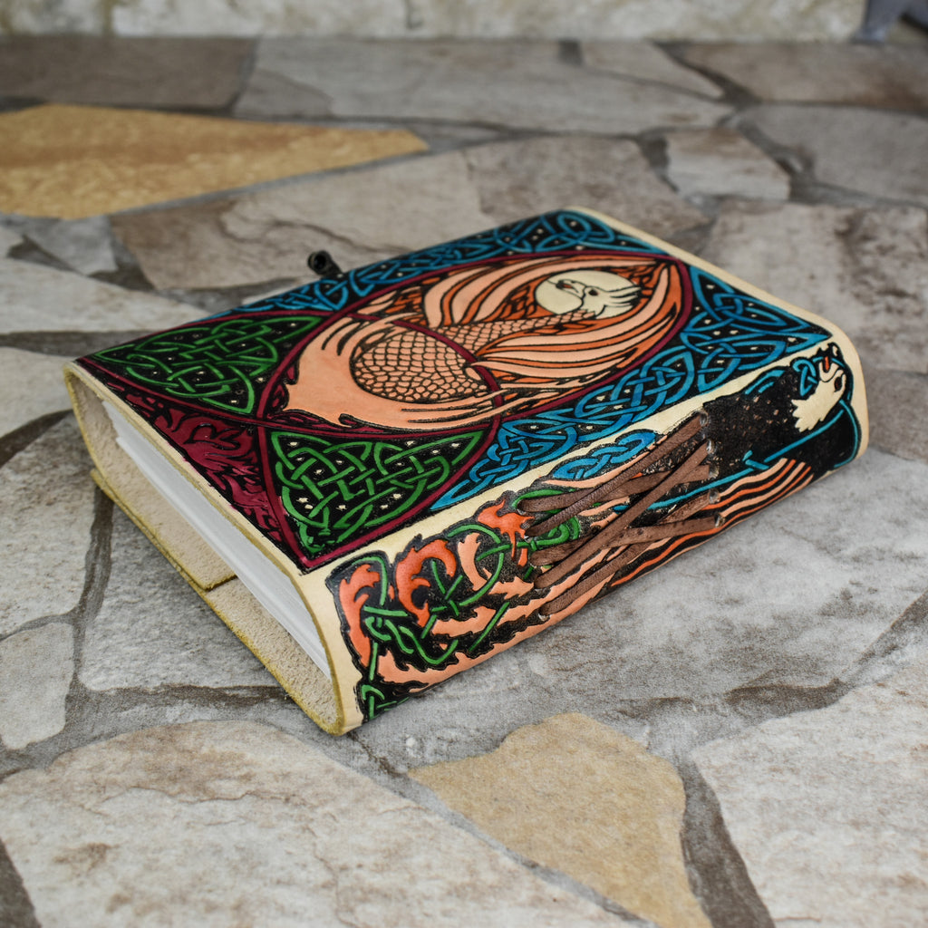 A hand-bound leather adventure journal with a full-grain goat leather cover featuring a Celtic inspired painted eagle design in brown, blue, and green.