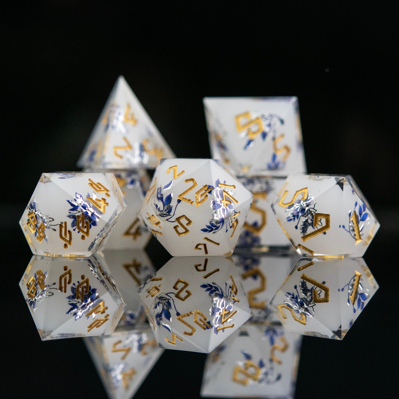 A set of white sharp-edged resin polyhedral dnd dice with hand-painted blue foral designs like a tea cup and blue numbers, reflecting on a black surface.