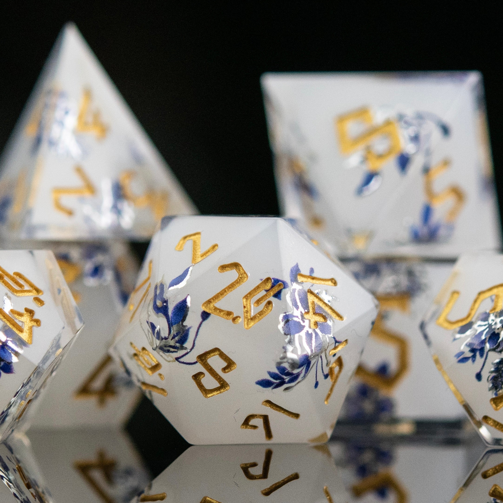 A set of white sharp-edged resin polyhedral dnd dice with hand-painted blue foral designs like a tea cup and blue numbers, reflecting on a black surface.