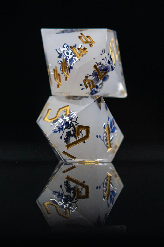 A set of white sharp-edged resin polyhedral dnd dice with hand-painted blue foral designs like a tea cup and blue numbers, reflecting on a black surface.