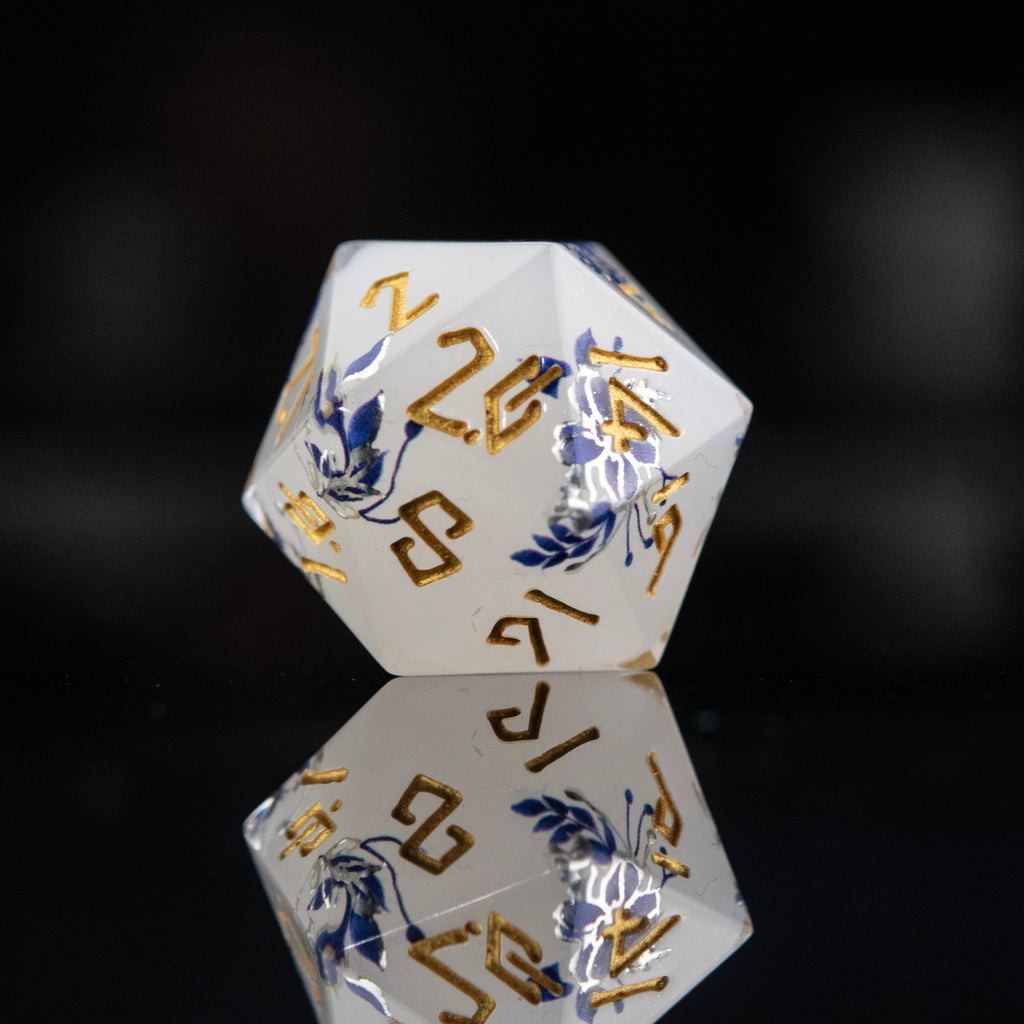 
A set of white sharp-edged resin polyhedral dnd dice with hand-painted blue foral designs like a tea cup and blue numbers, reflecting on a black surface.
