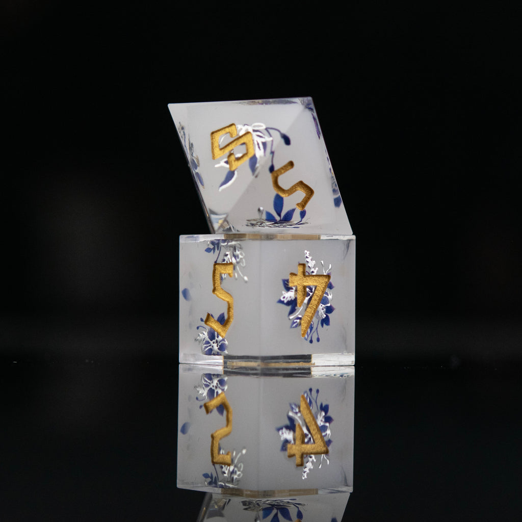 A set of white sharp-edged resin polyhedral dnd dice with hand-painted blue foral designs like a tea cup and blue numbers, reflecting on a black surface.