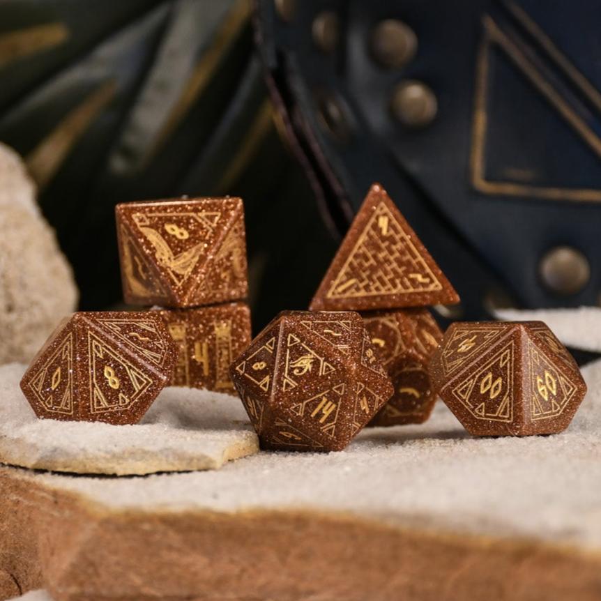 A set of hand-carved, gold-colored stone polyhedral dnd dice with Egyptian-inspired designs, displayed on a sandy surface.