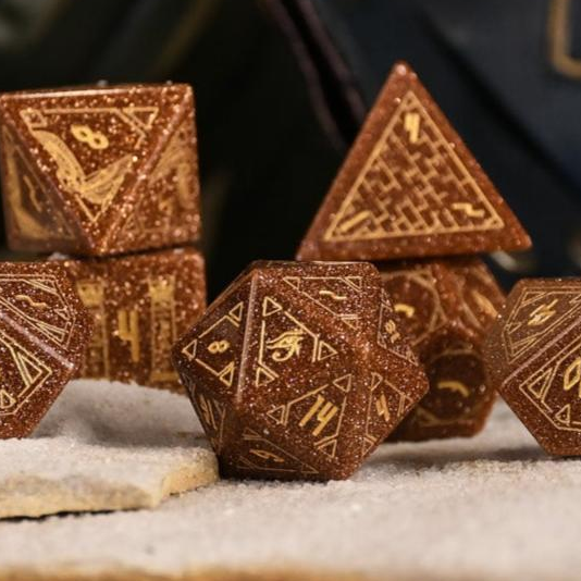 A set of hand-carved, gold-colored stone polyhedral dnd dice with Egyptian-inspired designs, displayed on a sandy surface.