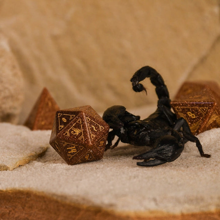 A set of hand-carved, gold-colored stone polyhedral dnd dice with Egyptian-inspired designs, displayed on a sandy surface.