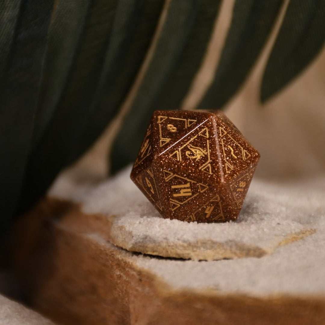 A set of hand-carved, gold-colored stone polyhedral dnd dice with Egyptian-inspired designs, displayed on a sandy surface.