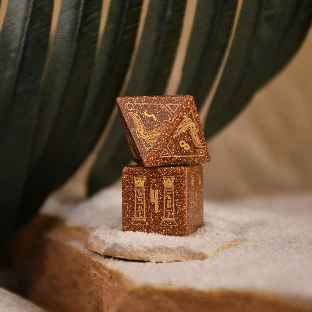A set of hand-carved, gold-colored stone polyhedral dnd dice with Egyptian-inspired designs, displayed on a sandy surface.