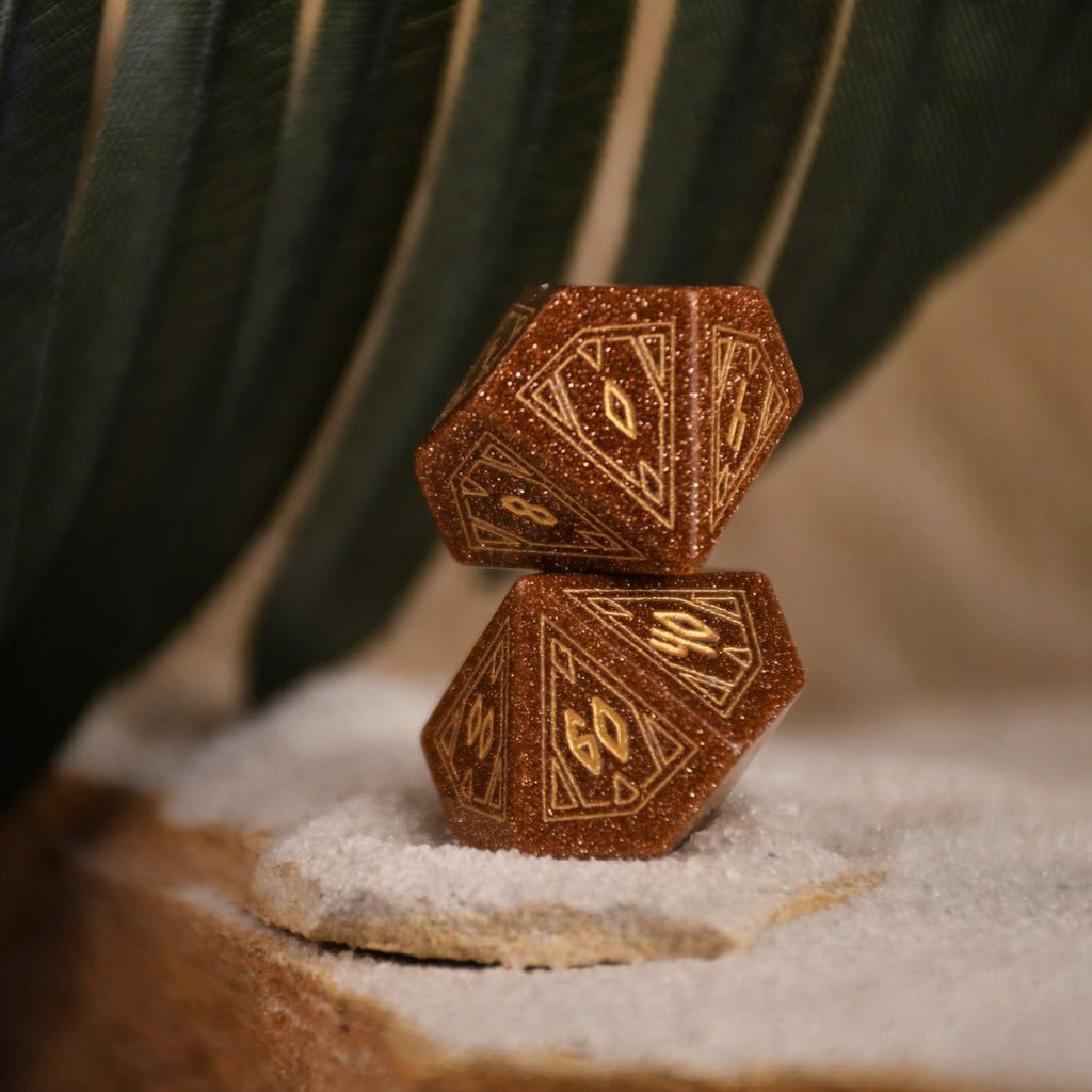 A set of hand-carved, gold-colored stone polyhedral dnd dice with Egyptian-inspired designs, displayed on a sandy surface.