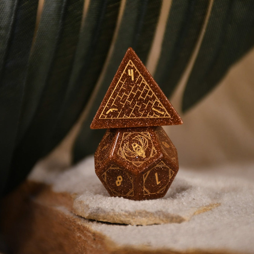 A set of hand-carved, gold-colored stone polyhedral dnd dice with Egyptian-inspired designs, displayed on a sandy surface.