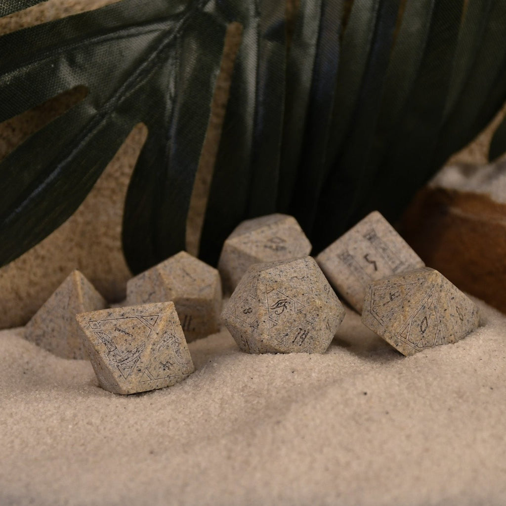 A set Egyptian Limestone Stone dnd dice with engraved numbers and Egyptian-inspired designs, displayed on a sandy surface.