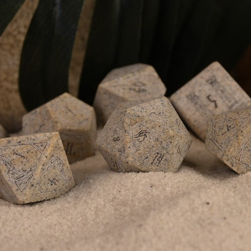 A set Egyptian Limestone Stone dnd dice with engraved numbers and Egyptian-inspired designs, displayed on a sandy surface.