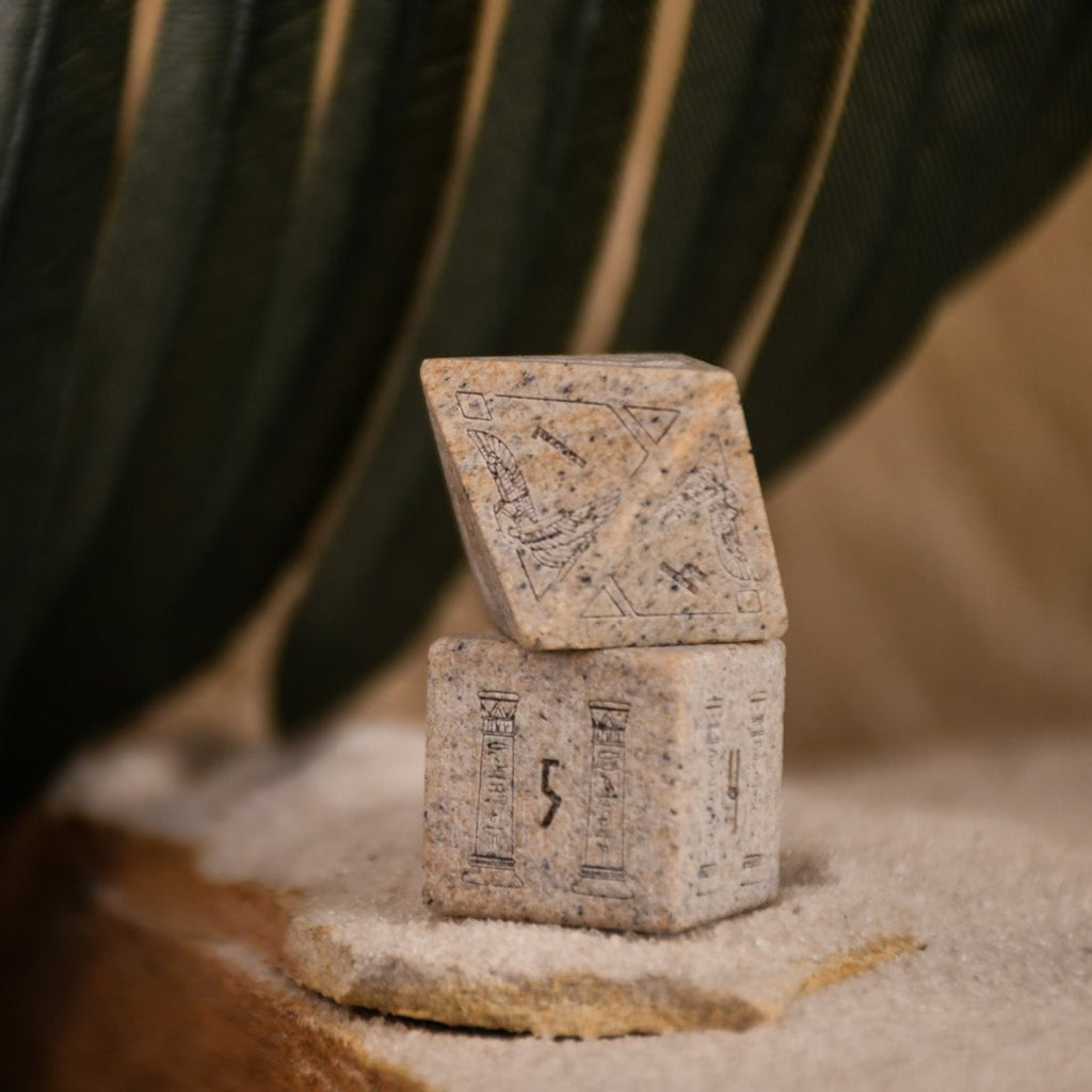 A set Egyptian Limestone Stone dnd dice with engraved numbers and Egyptian-inspired designs, displayed on a sandy surface.