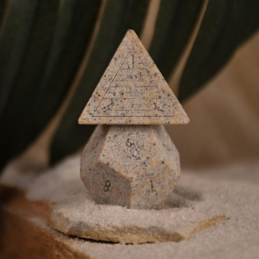 A set Egyptian Limestone Stone dnd dice with engraved numbers and Egyptian-inspired designs, displayed on a sandy surface.