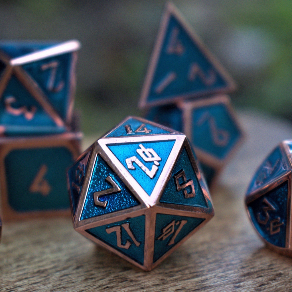 A set of metal dice in bronze and blue colors, including different shapes for a role-playing game. The set is displayed on a white surface with flowers in the background.