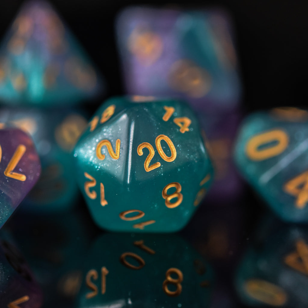 A set of acrylic ttrpg dice with engraved gold numbers, featuring a teal color with purple highlights, displayed on a reflective surface.