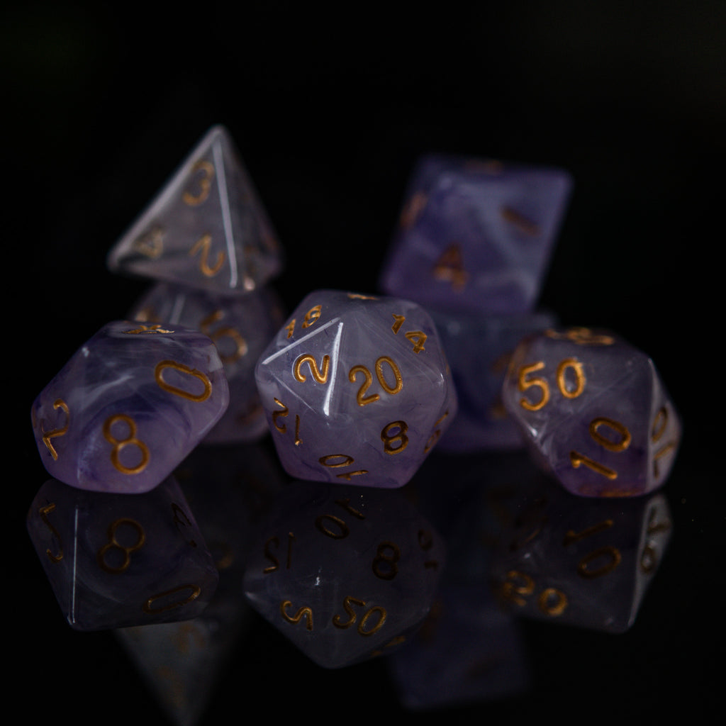 A set of creamy purple acrylic dice with golden engraved numbers, displayed on a reflective surface.