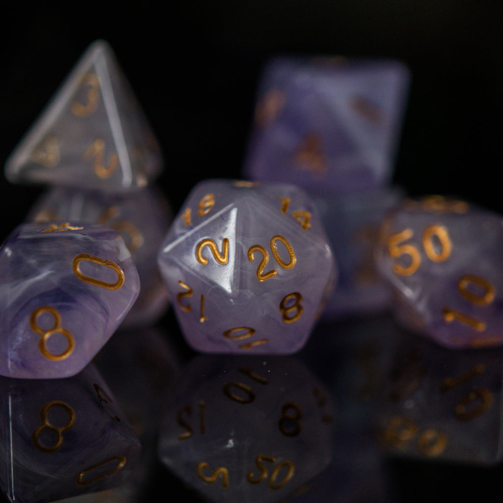 A set of creamy purple acrylic dice with golden engraved numbers, displayed on a reflective surface.