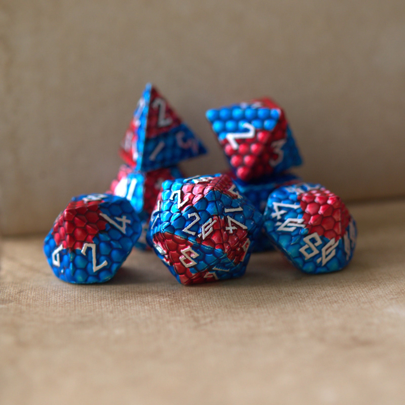 A set of metal dice with embossed silver numbers, featuring red and blue dragon egg patterns, displayed on a beige surface.