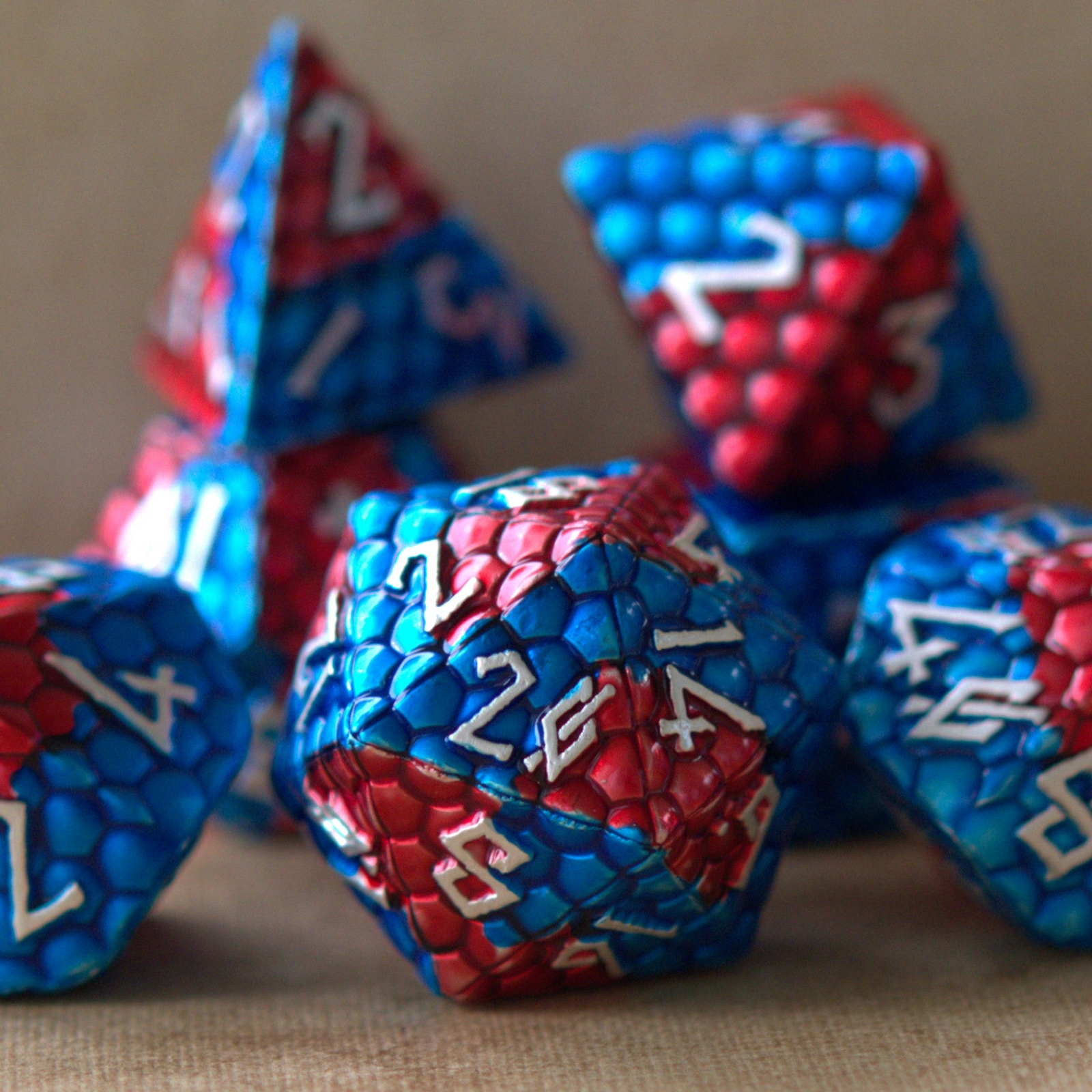A set of metal dice with embossed silver numbers, featuring red and blue dragon egg patterns, displayed on a beige surface.