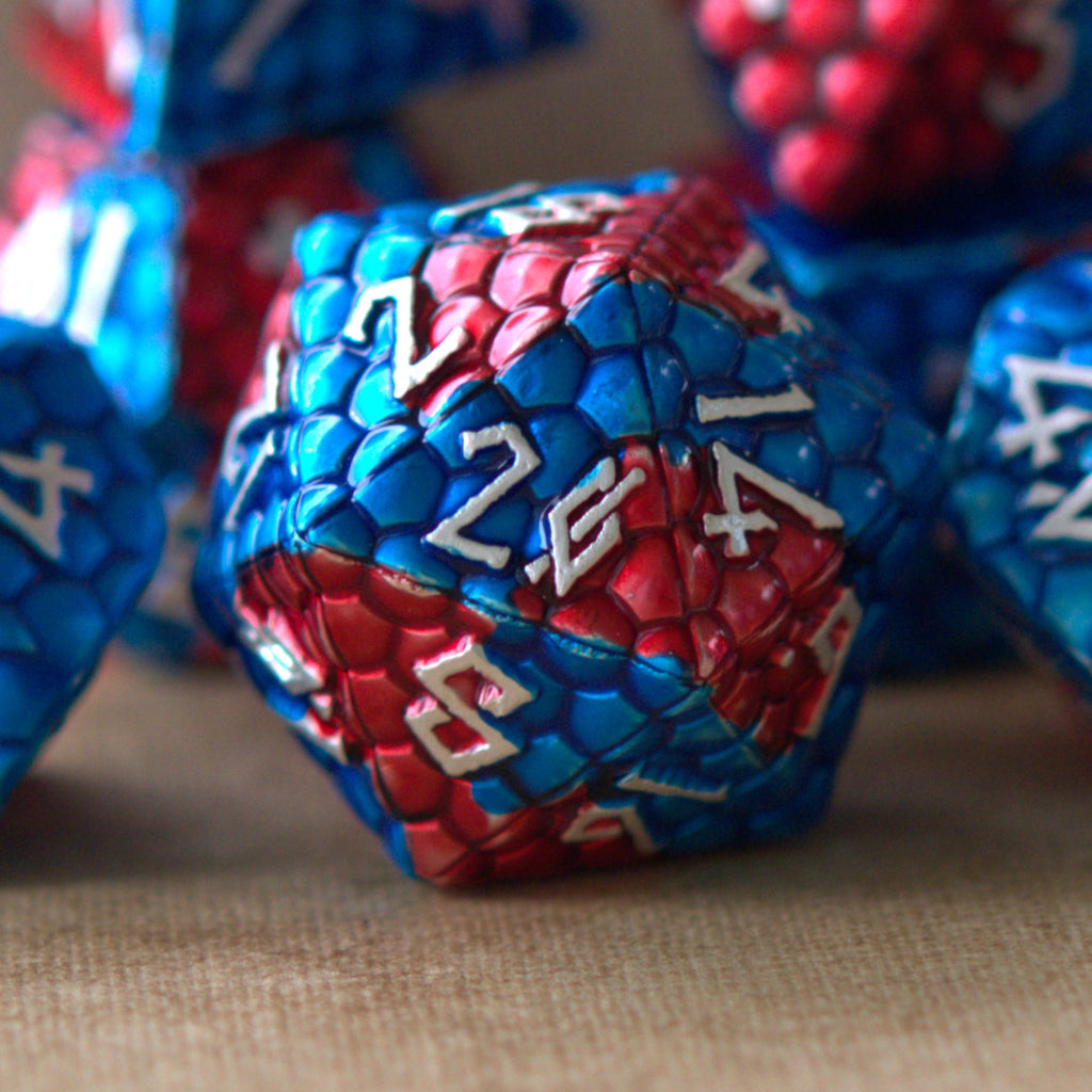 A set of metal dice with embossed silver numbers, featuring red and blue dragon egg patterns, displayed on a beige surface.