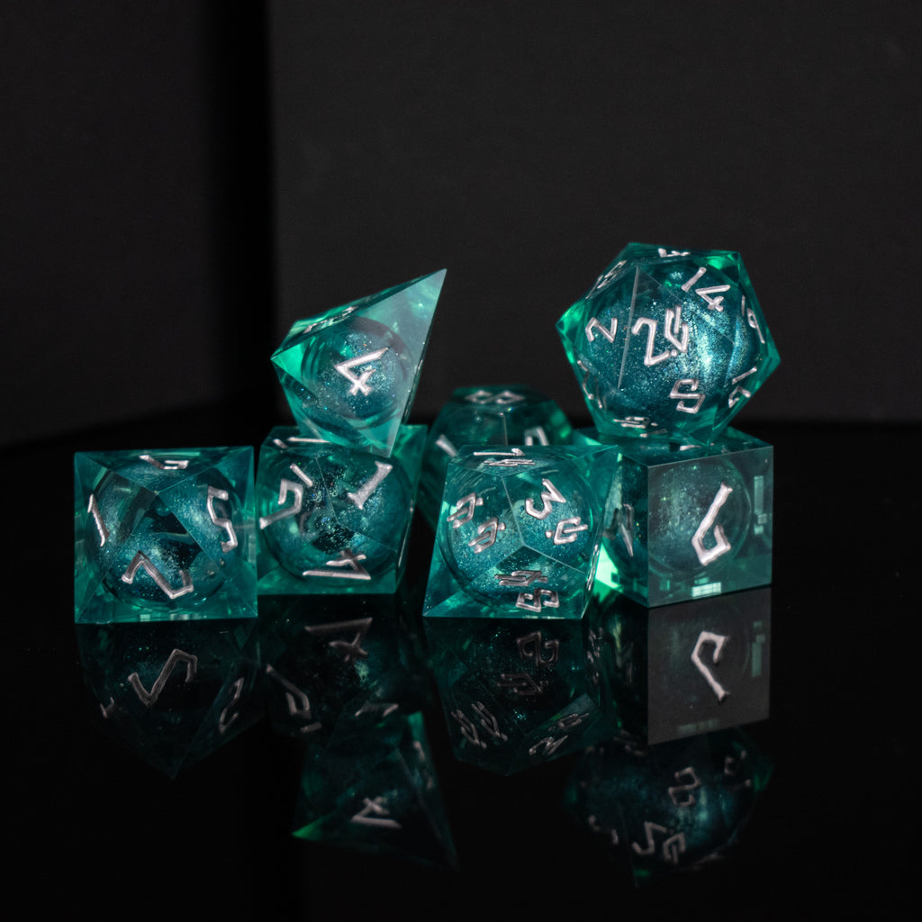 A set of teal resin dice with silver hand-painted numbers, including various shapes such as a d4, d6, d8, d10, d12, and d20, displayed on a reflective surface.