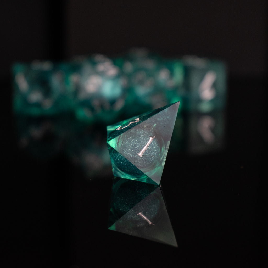 A set of teal resin dice with silver hand-painted numbers, including various shapes such as a d4, d6, d8, d10, d12, and d20, displayed on a reflective surface.
