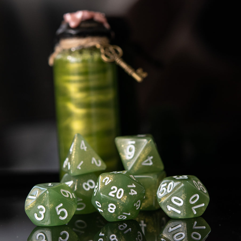 A set of green glittery acrylic dice with white numbering, displayed in front of a translucent container with a lid, against a dark background.