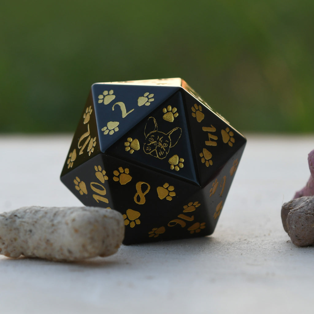 A black 30mm obsidian D20 dice with gold puppy paw prints and numbers, placed on a rock with a blurred natural background.