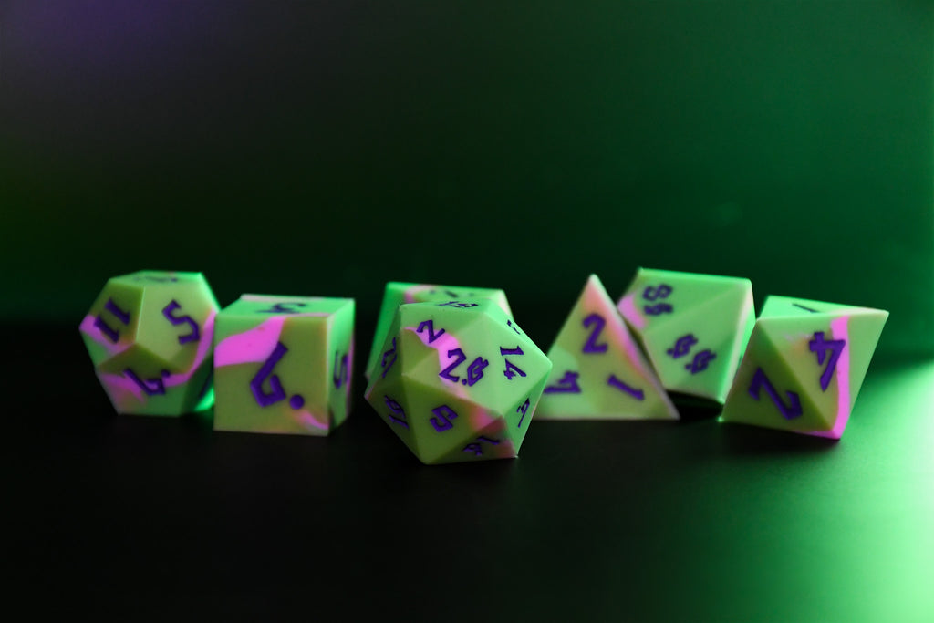 A set of silicone dice in various shapes including a D4, D6, D8, D10, D12, and D20, with green coloring and purple numbers. The dice are displayed against a dark background.